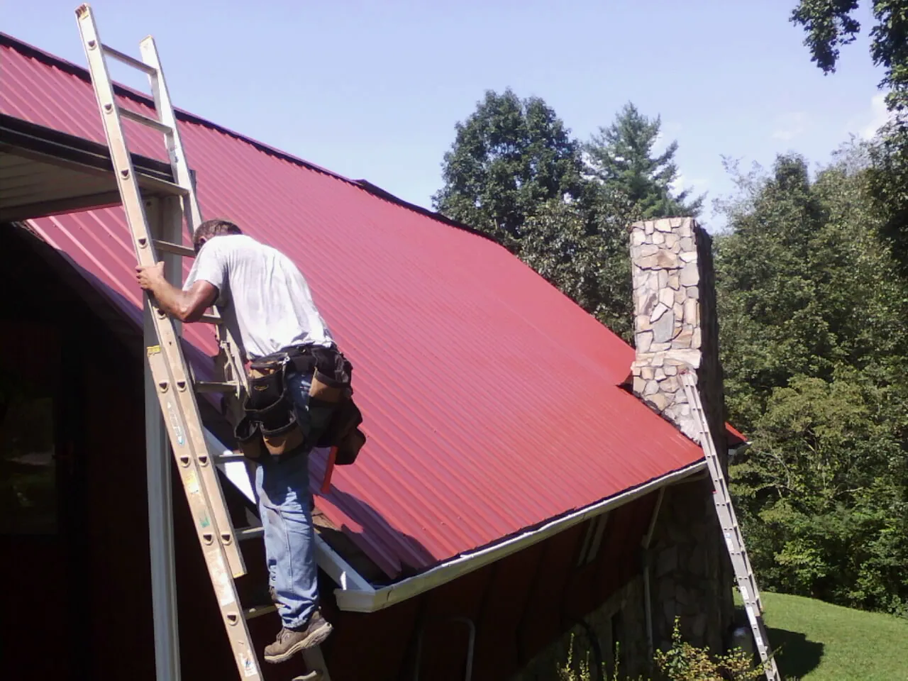 Roofer on a ladder beside a red metal roof and stone chimney — Soffit & Fascia Repair by Roofing Brothers in Camano Island